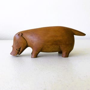 K&ouml;nnte beinhalten: Eine geschnitzte Holzfigur eines Schweins mit kleinem Schwanz. Das Schwein ist braun und hat einen glatten Abschluss und einen l&auml;chelnden Ausdruck. Die Figur ist ein dekorativer Artikel.