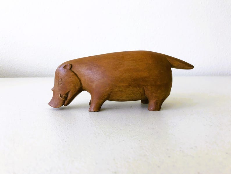 K&ouml;nnte beinhalten: Eine geschnitzte Holzfigur eines Schweins mit kleinem Schwanz. Das Schwein ist braun und hat einen glatten Abschluss und einen l&auml;chelnden Ausdruck. Die Figur ist ein dekorativer Artikel.