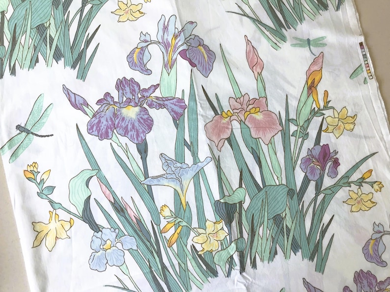 May include: White fabric with a floral pattern featuring irises in shades of purple, pink, and blue, along with yellow flowers and green foliage. The design includes dragonflies. The fabric is likely used for home decor or apparel.