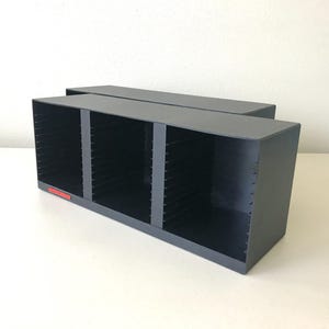 May include: A gray storage organizer with four compartments. The organizer has a rectangular shape and a red label on the front. The compartments are designed to hold items, likely for organization.