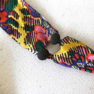 Op de afbeelding: Een kleurrijke stoffen ketting met een levendig patroon van rood, geel, groen en roze. De ketting is verbonden met twee zwarte, getextureerde, kogelvormige sluitingen. Het ontwerp is gedurfd en opvallend.