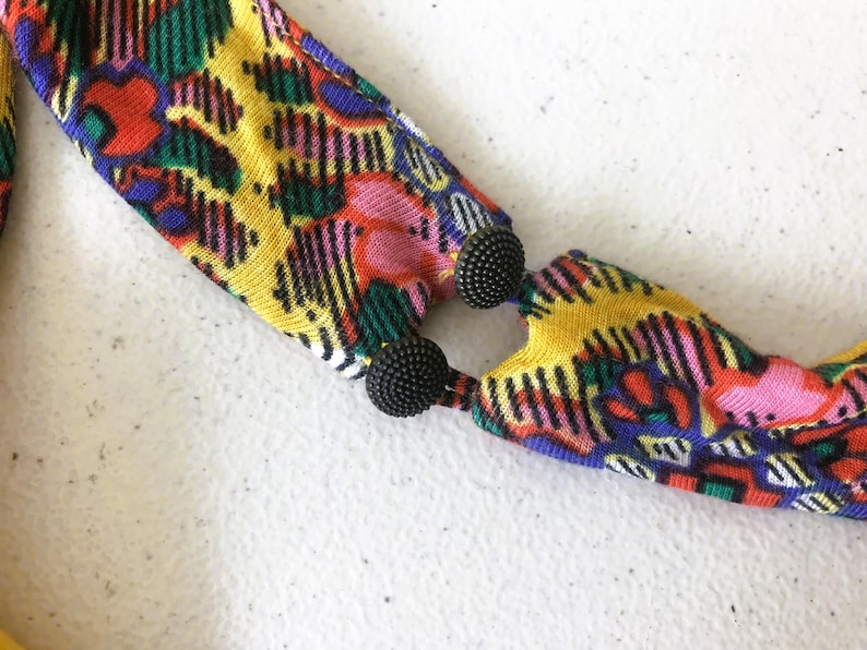 Op de afbeelding: Een kleurrijke stoffen ketting met een levendig patroon van rood, geel, groen en roze. De ketting is verbonden met twee zwarte, getextureerde, kogelvormige sluitingen. Het ontwerp is gedurfd en opvallend.