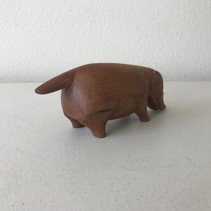 K&ouml;nnte beinhalten: Eine braune Holzskulptur eines Tieres, m&ouml;glicherweise eines Dackels, von hinten gesehen. Die Skulptur hat einen Schwanz und vier Beine. Sie steht auf einer wei&szlig;en Oberfl&auml;che, mit einer strukturierten wei&szlig;en Wand im Hintergrund.