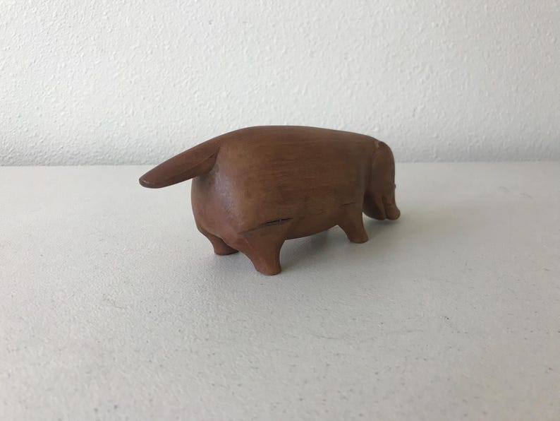 K&ouml;nnte beinhalten: Eine braune Holzskulptur eines Tieres, m&ouml;glicherweise eines Dackels, von hinten gesehen. Die Skulptur hat einen Schwanz und vier Beine. Sie steht auf einer wei&szlig;en Oberfl&auml;che, mit einer strukturierten wei&szlig;en Wand im Hintergrund.