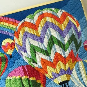 May include: A framed textile art piece featuring a vibrant scene of hot air balloons and a rainbow against a blue sky. The balloons are in various colors, including green, orange, pink, and purple. The artwork is framed in a gold-colored frame.