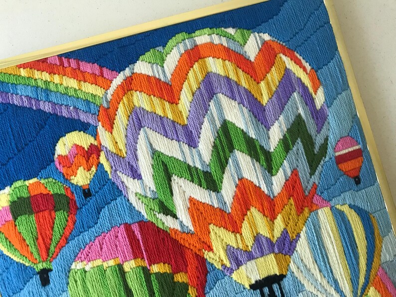 May include: A framed textile art piece featuring a vibrant scene of hot air balloons and a rainbow against a blue sky. The balloons are in various colors, including green, orange, pink, and purple. The artwork is framed in a gold-colored frame.