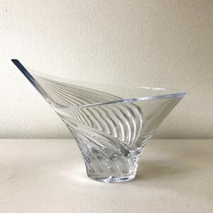 Pode incluir: Tigela decorativa de vidro transparente com um design assim&eacute;trico &uacute;nico. A tigela apresenta uma forma alargada, semelhante a uma asa, com linhas verticais texturizadas. O vidro transparente permite a passagem da luz, real&ccedil;ando os detalhes.