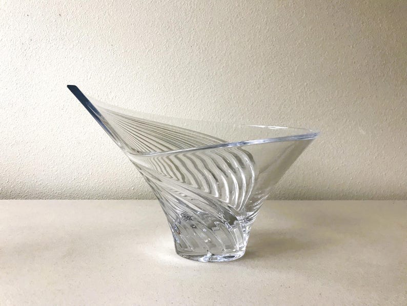 Pode incluir: Tigela decorativa de vidro transparente com um design assim&eacute;trico &uacute;nico. A tigela apresenta uma forma alargada, semelhante a uma asa, com linhas verticais texturizadas. O vidro transparente permite a passagem da luz, real&ccedil;ando os detalhes.