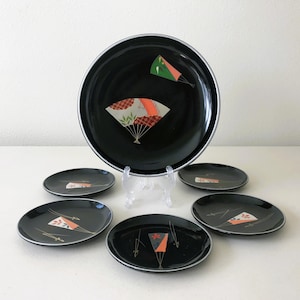 May include: Set of six black and gold Japanese-style plates with a fan design. The larger plate has a green and red fan design, while the smaller plates have a red and gold fan design.