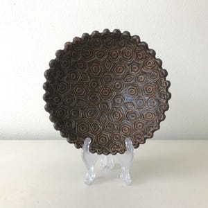 May include: A circular brown bowl featuring a repeating geometric pattern of dark brown hexagons and pentagons across its interior surface. The rim of the bowl has a scalloped, beaded edge, and the entire piece is displayed on a clear stand.