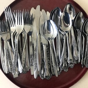 May include: A collection of assorted stainless steel silverware including forks, knives, and spoons with various decorative handle patterns, all resting on a round, dark red tray featuring ornate, cutout metal handles on each side.