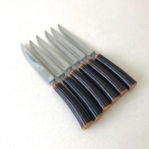 May include: A set of six steak knives with silver-toned stainless steel blades and dark brown, textured handles that resemble natural antler. The knives are arranged in a parallel, overlapping formation, showcasing the metallic blades and the distinct, rugged grip of the handles.