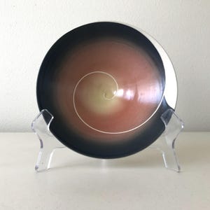 May include: A decorative ceramic bowl with a spiral design. The bowl features a gradient of colors, transitioning from a dark navy blue to a warm peach and yellow center. A white spiral detail is in the center of the bowl. The bowl is displayed on a clear acrylic stand.