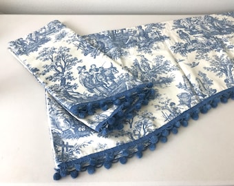 May include: A blue and white toile table runner and matching pillow cover. The fabric features a pastoral scene with figures, trees, and buildings. The edges of the runner and pillow cover are trimmed with blue pom-poms.