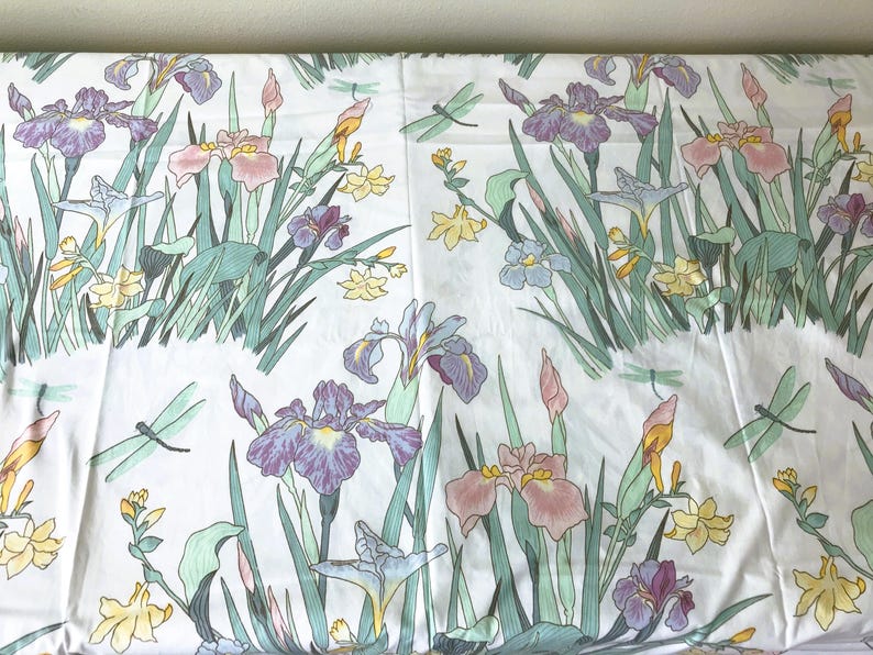 May include: White fabric with a floral pattern featuring purple, pink, and yellow irises, green leaves, and light green dragonflies. The design is repeated across the fabric, creating a garden-like scene.