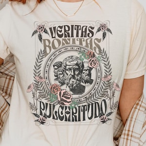 May include: Ivory t-shirt with a graphic print of three men's faces in a circle with the text "VERITAS BONITAS PULCHRITUDO" and floral accents.