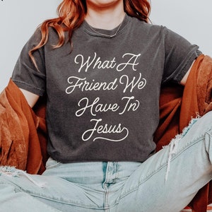 Könnte beinhalten: Eine Frau trägt ein dunkelgraues T-Shirt mit dem Text "What a Friend We Have In Jesus" in weißen Buchstaben. Sie sitzt auf einer rot-weiß karierten Oberfläche und trägt blaue Jeans und eine braune Jacke.