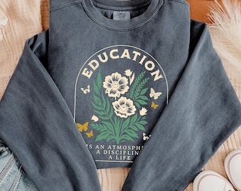 May include: A denim blue sweatshirt with the word "EDUCATION" arched above a floral design. The design includes white flowers, green leaves, and yellow butterflies. The text "IS AN ATMOSPHERE A DISCIPLINE A LIFE" is below the floral design. The word "denim" is at the bottom.