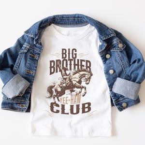 May include: White t-shirt featuring a brown graphic of a cowboy on a horse, with the text "BIG BROTHER YEE-HAW CLUB." A blue denim jacket is draped over the shirt.