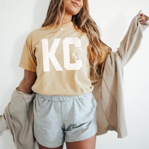 May include: A mustard yellow t-shirt with the letters "KC" in white, along with a football graphic. The person is wearing gray shorts and a light brown jacket. The word "butter" is in the bottom right corner.
