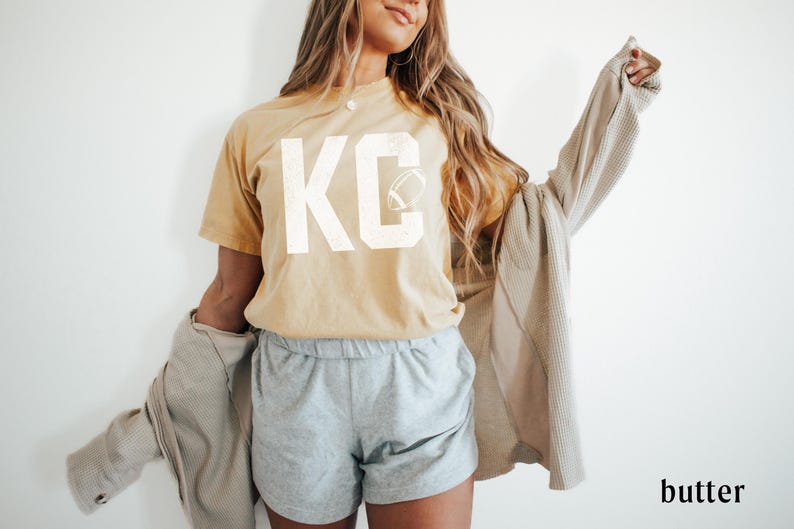 May include: A mustard yellow t-shirt with the letters "KC" in white, along with a football graphic. The person is wearing gray shorts and a light brown jacket. The word "butter" is in the bottom right corner.
