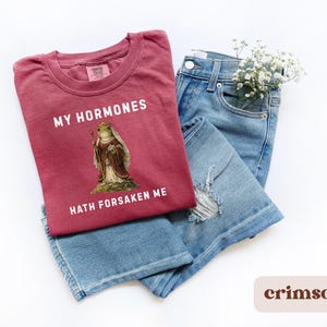 May include: Crimson t-shirt featuring the text "MY HORMONES HATH FORSAKEN ME" and a frog illustration in royal clothing. The shirt is laid on blue denim jeans. A small bunch of white flowers is placed in the jeans pocket.