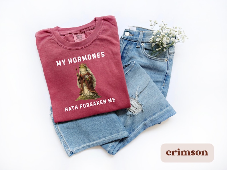 May include: Crimson t-shirt featuring the text "MY HORMONES HATH FORSAKEN ME" and a frog illustration in royal clothing. The shirt is laid on blue denim jeans. A small bunch of white flowers is placed in the jeans pocket.