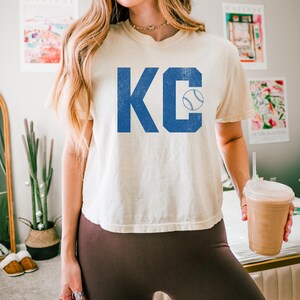May include: Cream-coloured t-shirt displaying the letters "KC" in blue, with a baseball graphic. The shirt has a loose fit and short sleeves. The word "ivory" is printed at the bottom of the image.