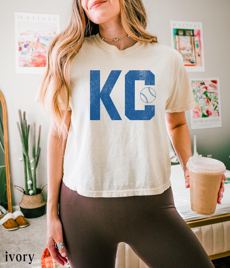 May include: Cream-coloured t-shirt displaying the letters "KC" in blue, with a baseball graphic. The shirt has a loose fit and short sleeves. The word "ivory" is printed at the bottom of the image.