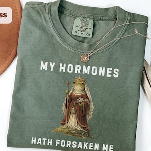 Könnte beinhalten: Moosgrünes T-Shirt mit dem Text "MY HORMONES HATH FORSAKEN ME" und einer Illustration einer königlichen Frosch. Eine goldene Halskette mit Anhänger liegt auf dem Shirt. Das Shirt ist gefaltet, mit einem braunen Hut und einer getrockneten Pflanze im Hintergrund.