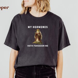 May include: Dark grey t-shirt featuring the text "MY HORMONES HATH FORSAKEN ME" above and below a vintage illustration of a frog in royal clothing. The shirt is a relaxed, casual style.