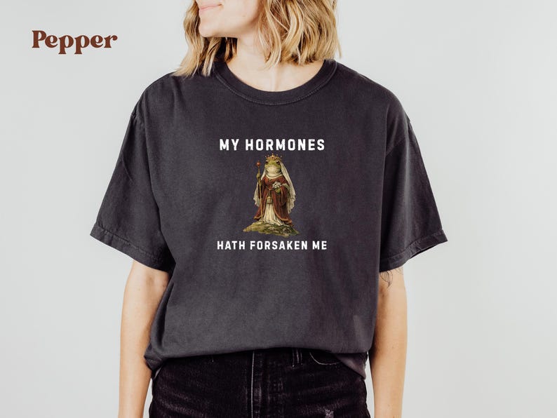 May include: Dark grey t-shirt featuring the text "MY HORMONES HATH FORSAKEN ME" above and below a vintage illustration of a frog in royal clothing. The shirt is a relaxed, casual style.