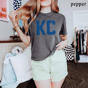 May include: A grey t-shirt featuring the blue letters "KC" and a baseball graphic. The shirt is worn with light green shorts. The word "pepper" is in the top right corner.
