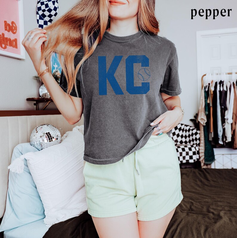 May include: A grey t-shirt featuring the blue letters "KC" and a baseball graphic. The shirt is worn with light green shorts. The word "pepper" is in the top right corner.
