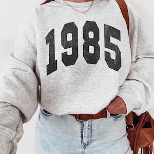 May include: A grey sweatshirt with the year 1985 printed in black on the front. The sweatshirt is being worn by a person wearing blue jeans and a brown belt.