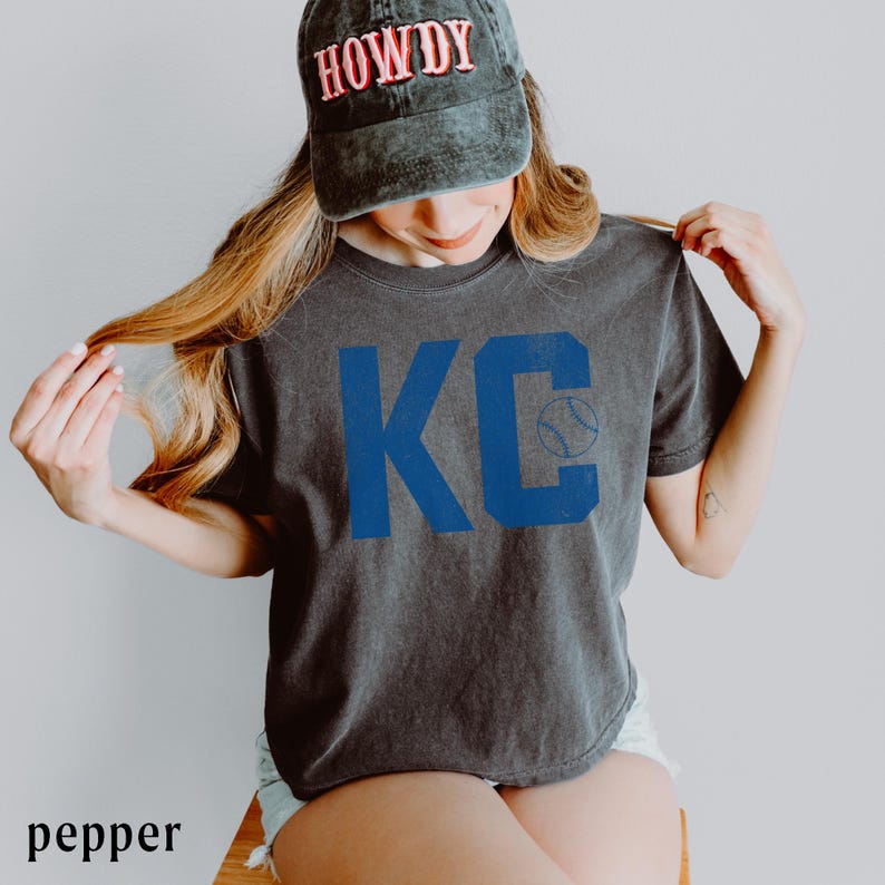 May include: A charcoal grey t-shirt featuring the blue letters "KC" and a baseball graphic. The person is wearing a faded black baseball cap with the word "HOWDY" in pink letters.