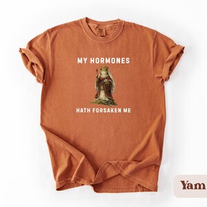 May include: A terracotta t-shirt with the text "MY HORMONES HATH FORSAKEN ME" in white. The shirt features a vintage illustration of a frog wearing a crown and robes. The tee shirt is a comfortable, casual style.