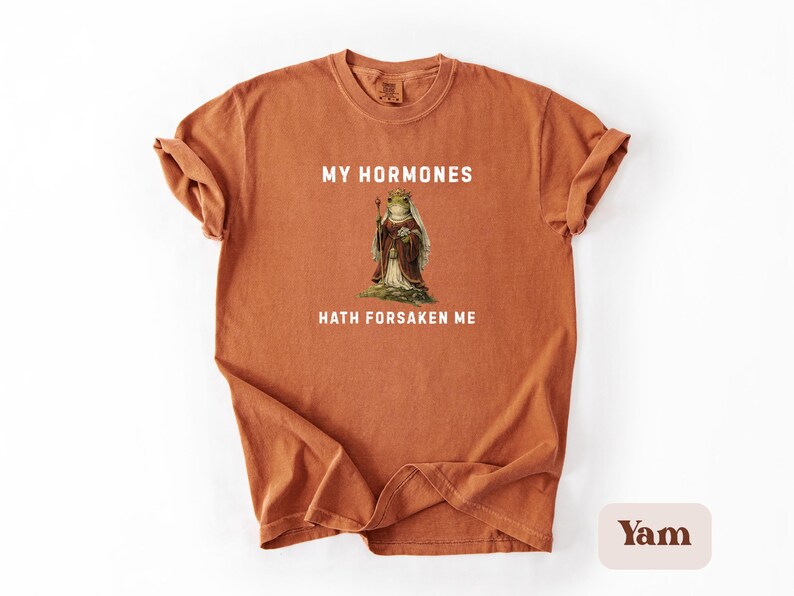 May include: A terracotta t-shirt with the text "MY HORMONES HATH FORSAKEN ME" in white. The shirt features a vintage illustration of a frog wearing a crown and robes. The tee shirt is a comfortable, casual style.