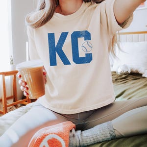 May include: An ivory-coloured t-shirt featuring the blue letters "KC" and a baseball graphic. The person is holding a beverage in a clear plastic cup. The person is wearing grey leggings and orange slippers.