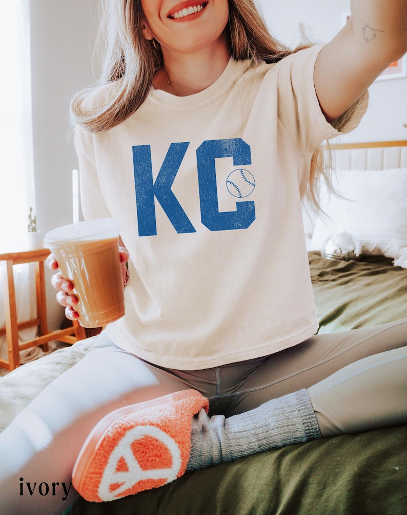 May include: An ivory-coloured t-shirt featuring the blue letters "KC" and a baseball graphic. The person is holding a beverage in a clear plastic cup. The person is wearing grey leggings and orange slippers.