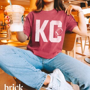 May include: Brick-colored t-shirt with the letters "KC" in white, featuring a football graphic. The person is wearing light blue jeans and white sneakers. The person is holding a Starbucks drink.