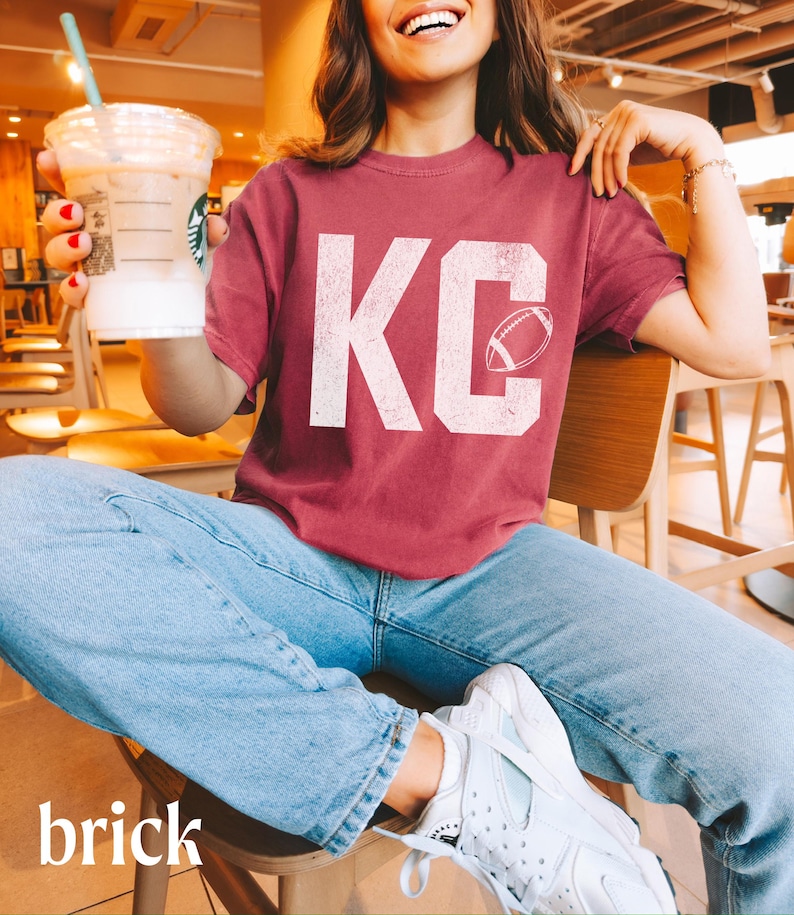 May include: Brick-colored t-shirt with the letters "KC" in white, featuring a football graphic. The person is wearing light blue jeans and white sneakers. The person is holding a Starbucks drink.