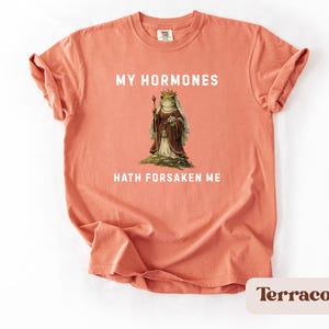 May include: A terracotta-coloured t-shirt with the text "MY HORMONES HATH FORSAKEN ME" in white. The shirt features a vintage illustration of a frog in royal attire. The t-shirt is a comfortable, casual style.