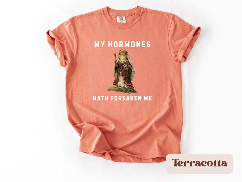 May include: A terracotta-coloured t-shirt with the text "MY HORMONES HATH FORSAKEN ME" in white. The shirt features a vintage illustration of a frog in royal attire. The t-shirt is a comfortable, casual style.
