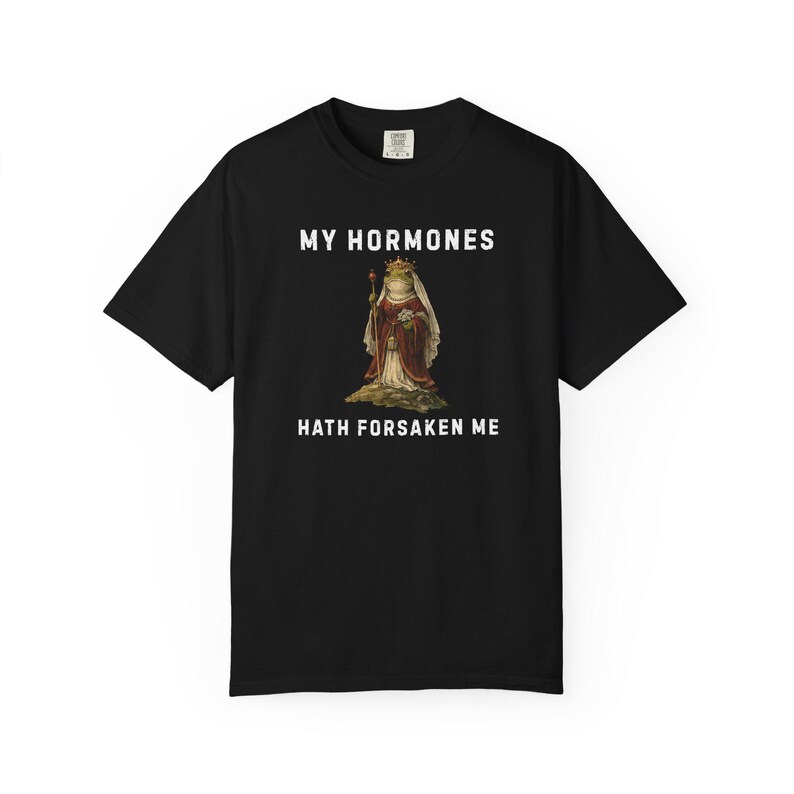 May include: Black t-shirt with the text "MY HORMONES HATH FORSAKEN ME" in white. The shirt features a graphic of a frog wearing a crown and royal attire, holding a staff. The t-shirt is a crew neck style.