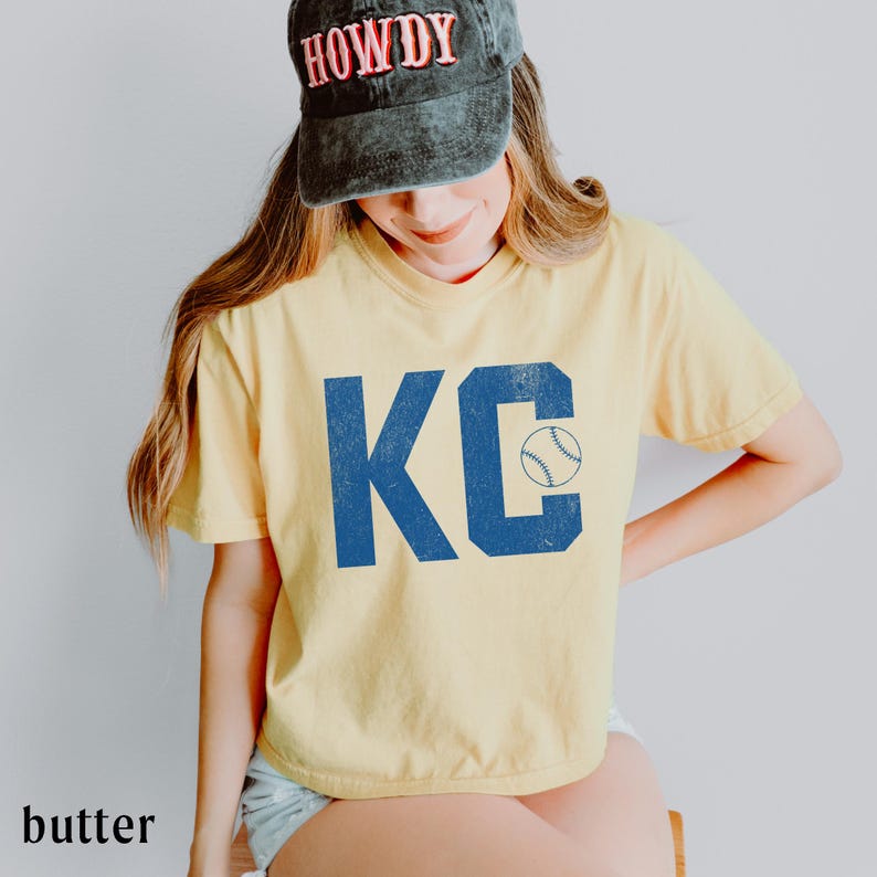 May include: A pale yellow t-shirt featuring the blue letters "KC" and a baseball graphic. The shirt is worn with a black baseball cap with the word "HOWDY" in red. The t-shirt has a relaxed fit.