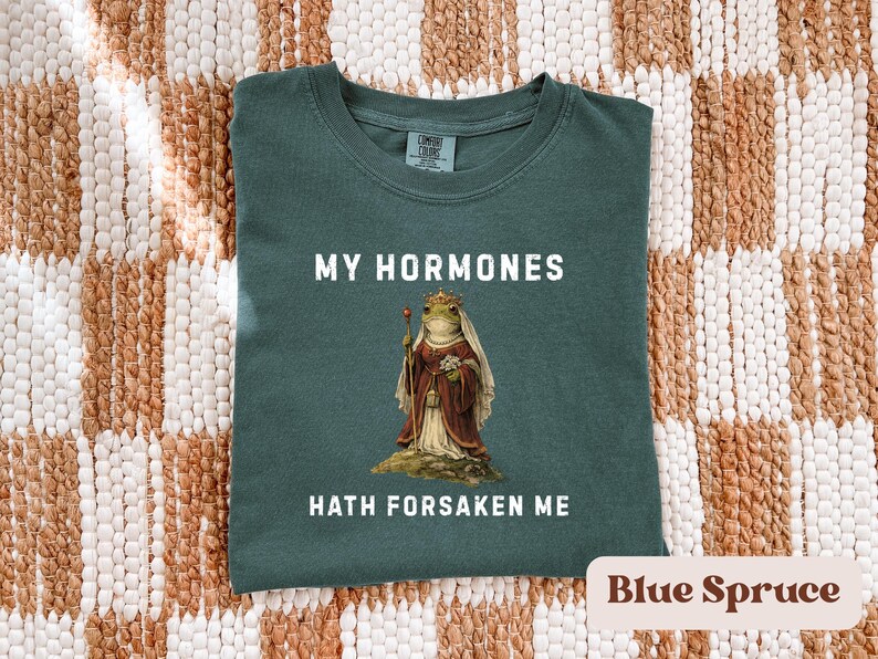 May include: A blue spruce t-shirt with the text "MY HORMONES HATH FORSAKEN ME" in white. The shirt features a regal frog illustration wearing a crown and holding a staff. The shirt is folded on a woven brown and white surface.