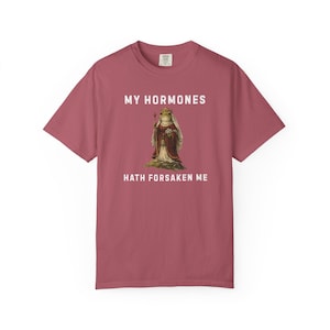 May include: A maroon t-shirt featuring the text "MY HORMONES HATH FORSAKEN ME" above and below a vintage illustration of a frog in royal clothing. The frog is wearing a crown and holding a staff.