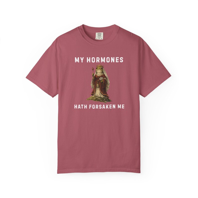 May include: A maroon t-shirt featuring the text "MY HORMONES HATH FORSAKEN ME" above and below a vintage illustration of a frog in royal clothing. The frog is wearing a crown and holding a staff.