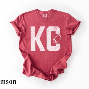 May include: Crimson-colored t-shirt with the letters "KC" in white, featuring a football graphic. The shirt has rolled-up sleeves and a crew neck. The word "crimson" is printed at the bottom of the image.